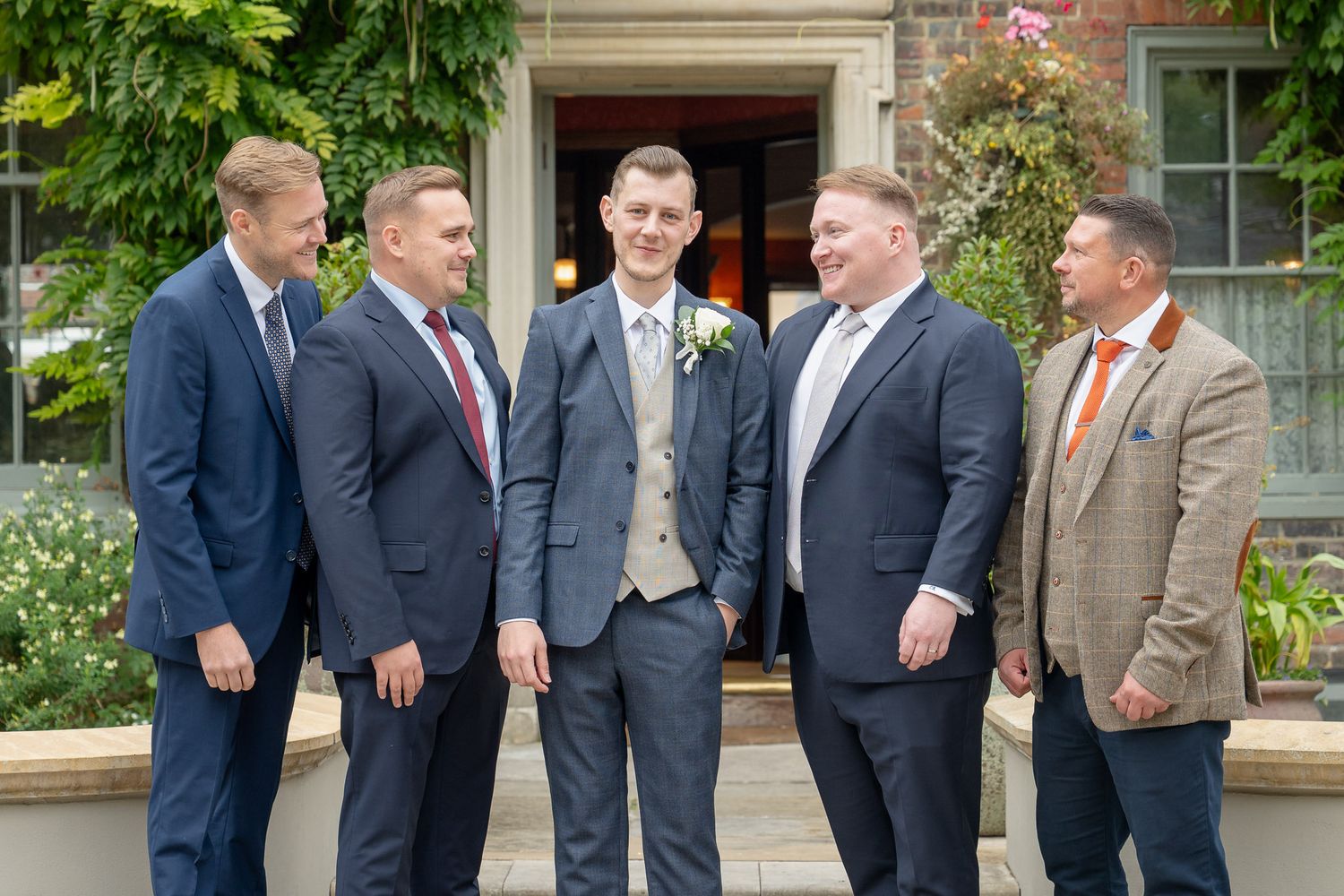 A group in formal attire stands together outdoors in front of a building with lush greenery and flowers.