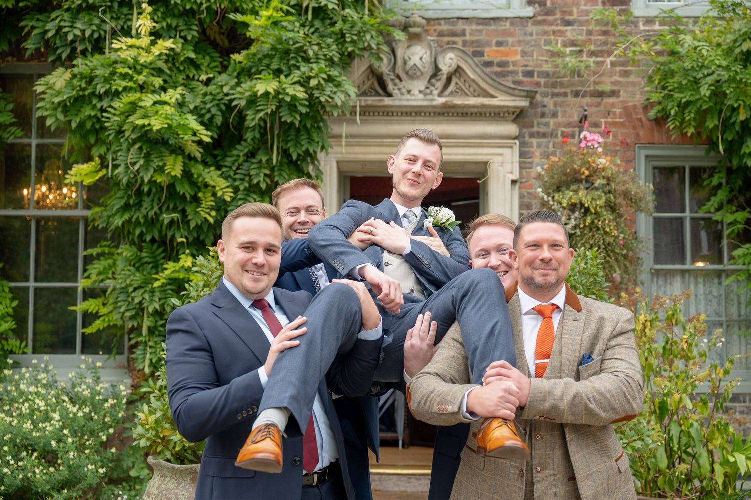 A group of groomsmen in dark suits pose playfully with beer bottles outside a brick building.