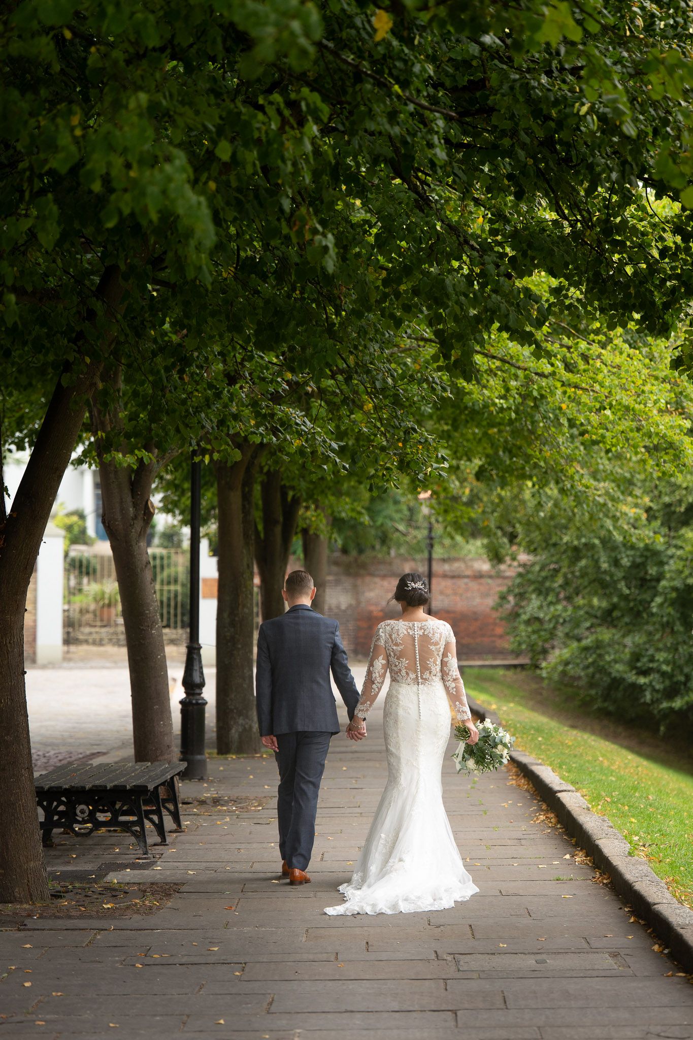 A couple in wedding attire walks along a tree-lined path in a romantic outdoor setting.