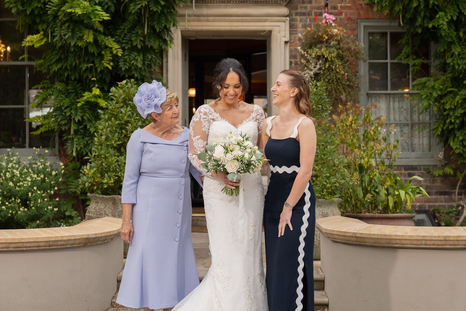 A group poses together in formal attire outside a brick building with lush greenery.