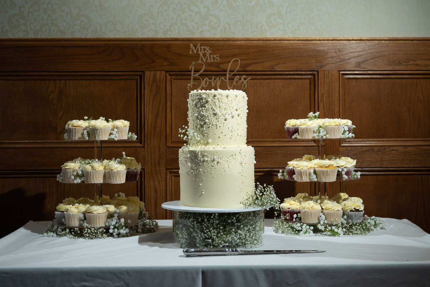 A white tiered wedding cake with delicate floral decorations displayed on a table with matching cupcakes and greenery.