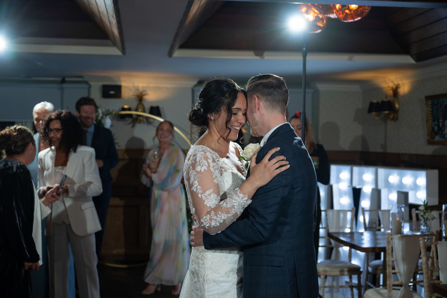 A couple shares their first dance as newlyweds in a dimly lit reception venue with guests watching in the background.