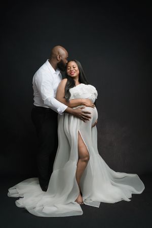 A couple shares an intimate embrace wearing white against a dark studio backdrop in a romantic photo series.