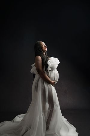 Elegant maternity portrait featuring flowing white gown against dark background in dramatic studio lighting.