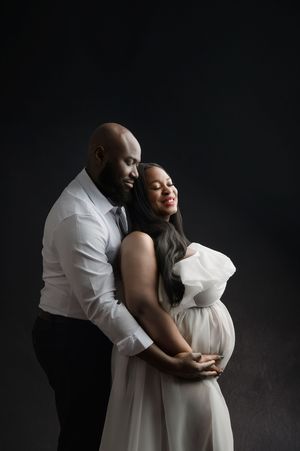 Couple in white clothing embraces lovingly during intimate maternity photoshoot against dark background.