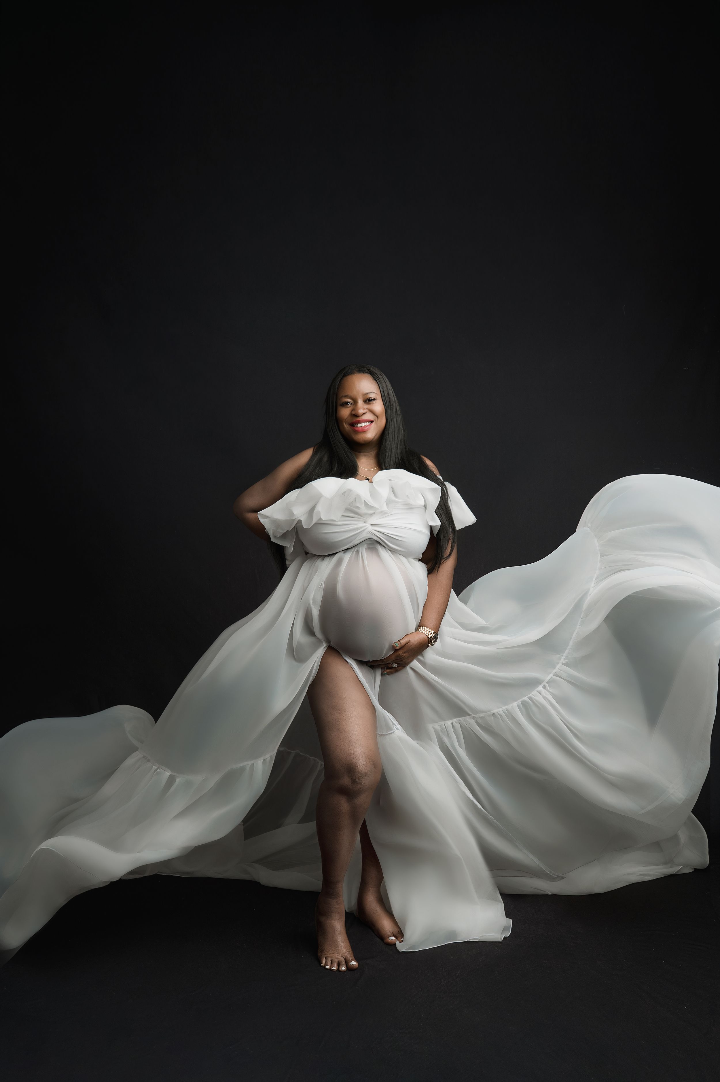 Elegant maternity portrait with flowing white fabric against dark background creating dramatic movement and contrast.