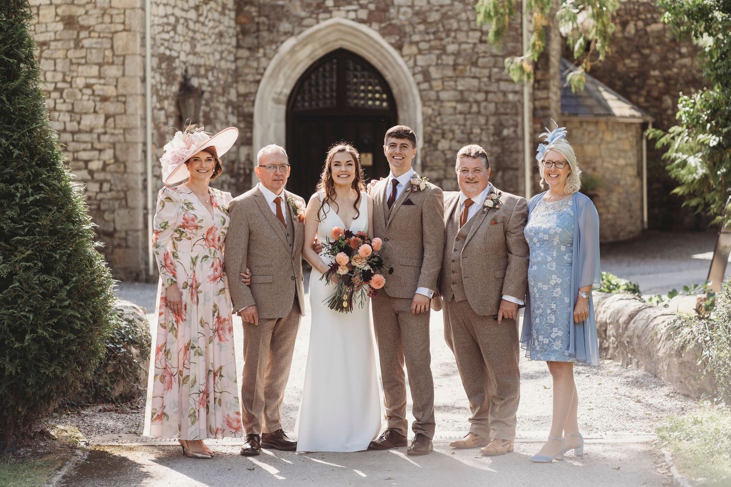 Bride and groom standing with their parents outside a stone building in South Wales, formal family wedding portrait in natural light.