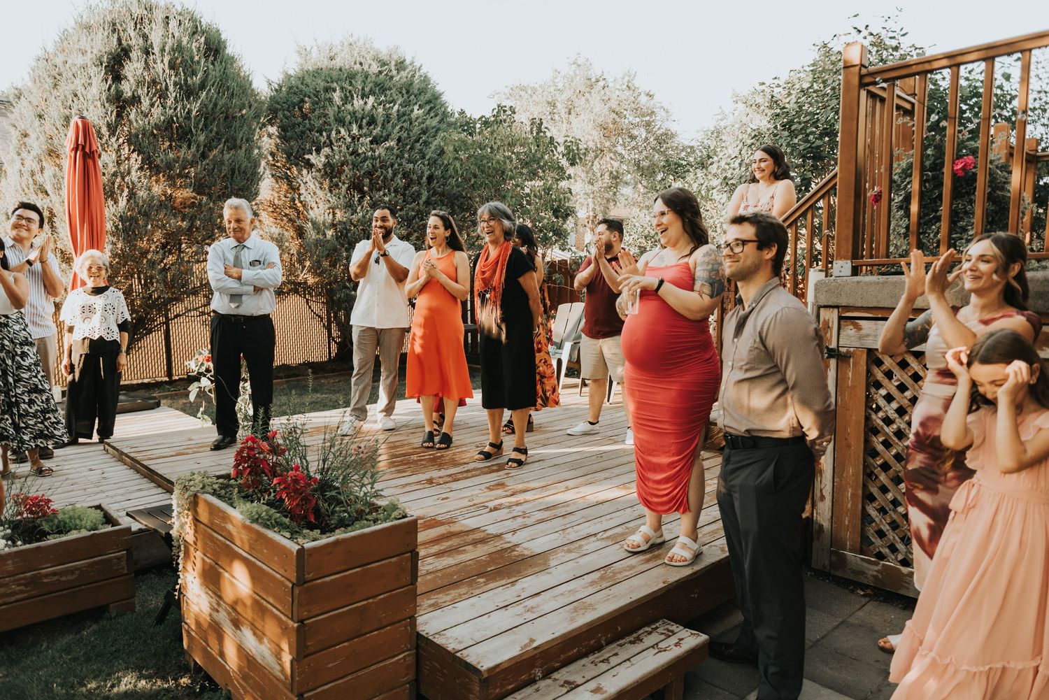 A group of guests gather on a wooden deck during an outdoor celebration with planters and decorative elements.