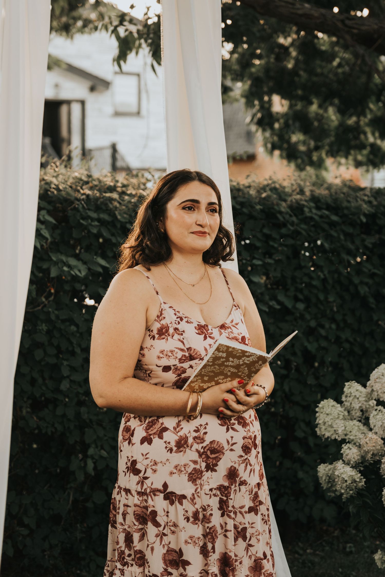 Person in a floral dress reading a book outdoors near white pillars and greenery on a sunny day.