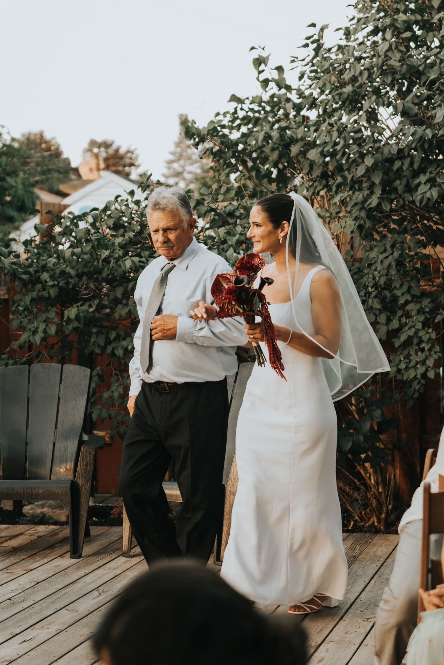 A couple in wedding attire shares a tender moment in front of a wooden fence and greenery during sunset.