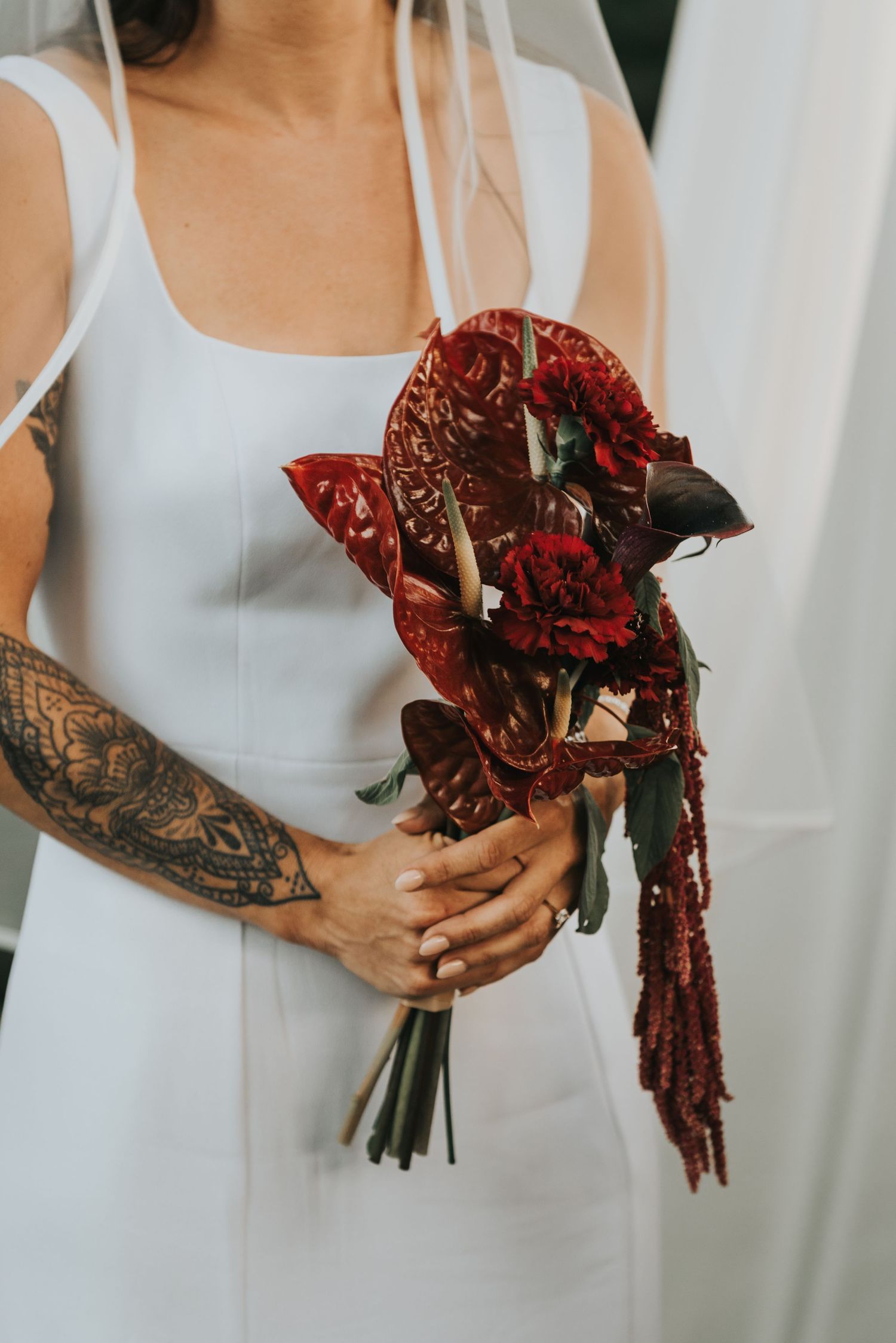 Bridesmaids in white dresses hold deep red rose bouquets with flowing burgundy ribbons against a light background.