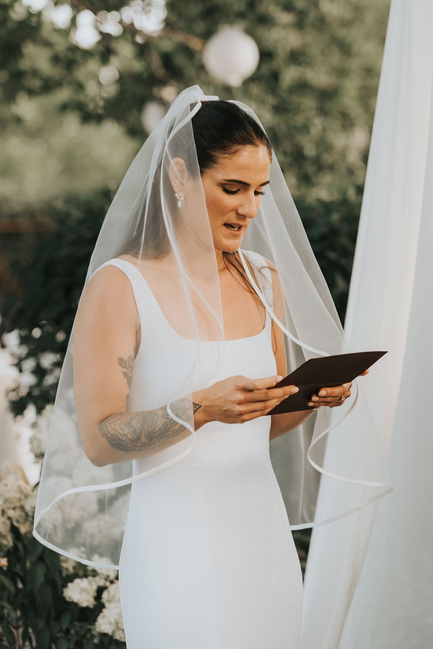 A bride in a white wedding dress and veil reads a letter during an emotional first look moment outdoors.