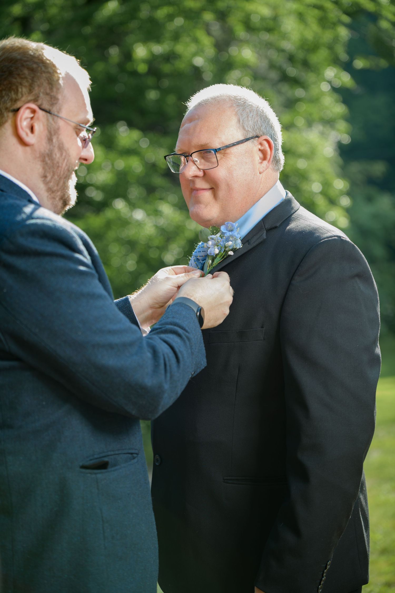 Man pinning a blue boutonniere on another mans suit lapel outdoors at a wedding ceremony.