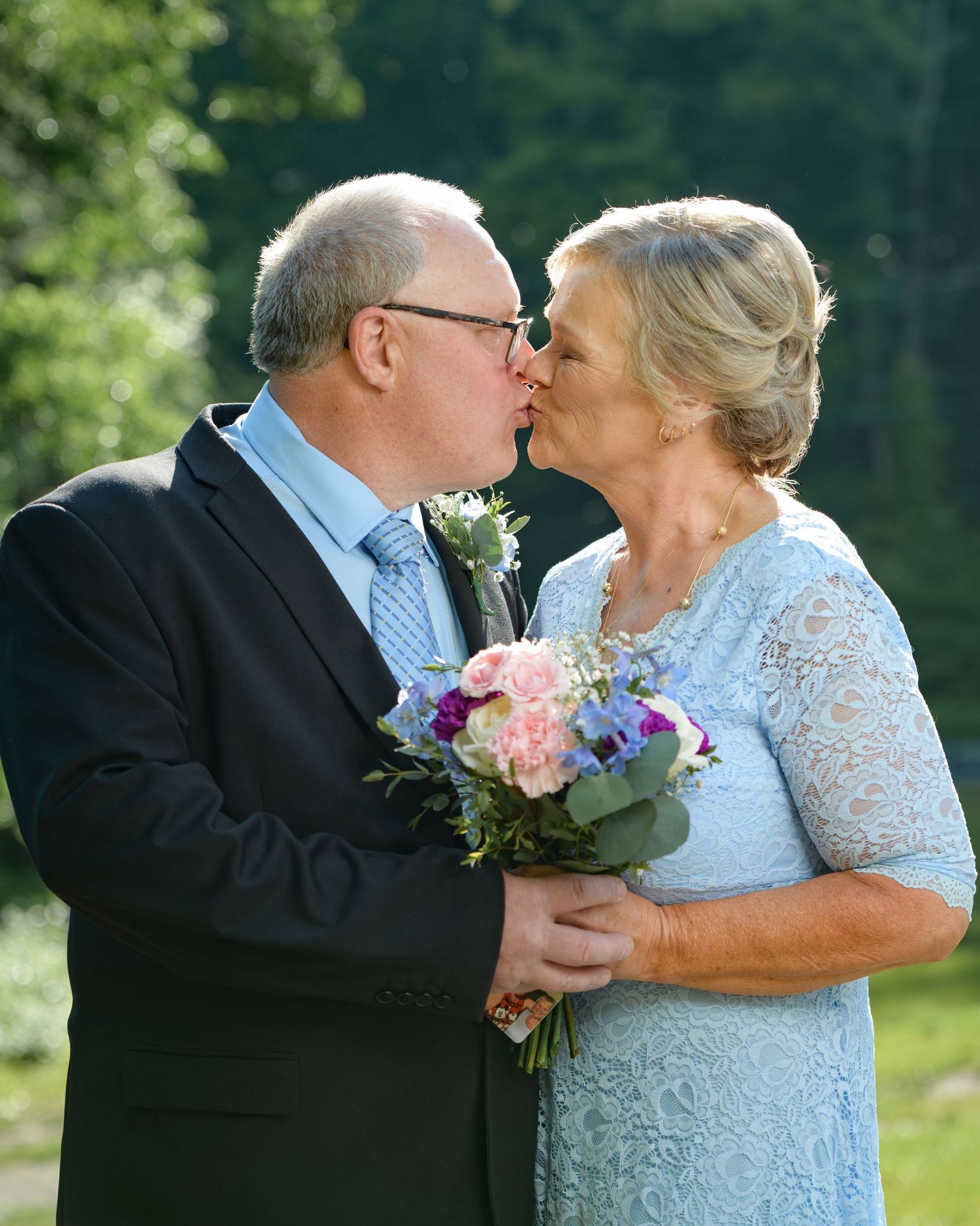 Older couple sharing a kiss while holding a colorful bridal bouquet at an outdoor wedding.