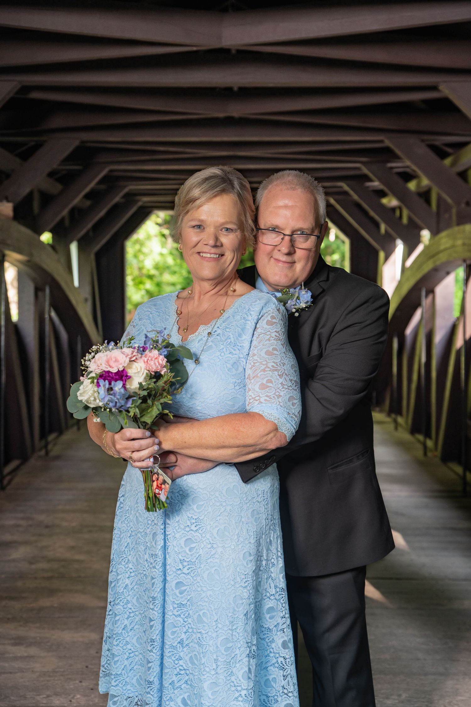 Smiling couple embracing inside a covered wooden bridge, woman holding a colorful wedding bouquet.