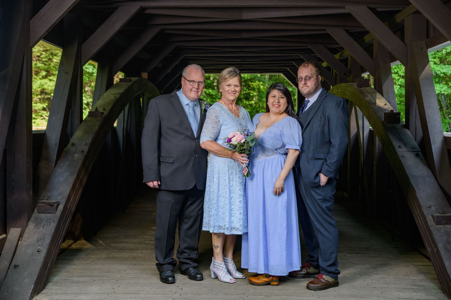 Group of four people posing together inside a rustic wooden covered bridge at a wedding.