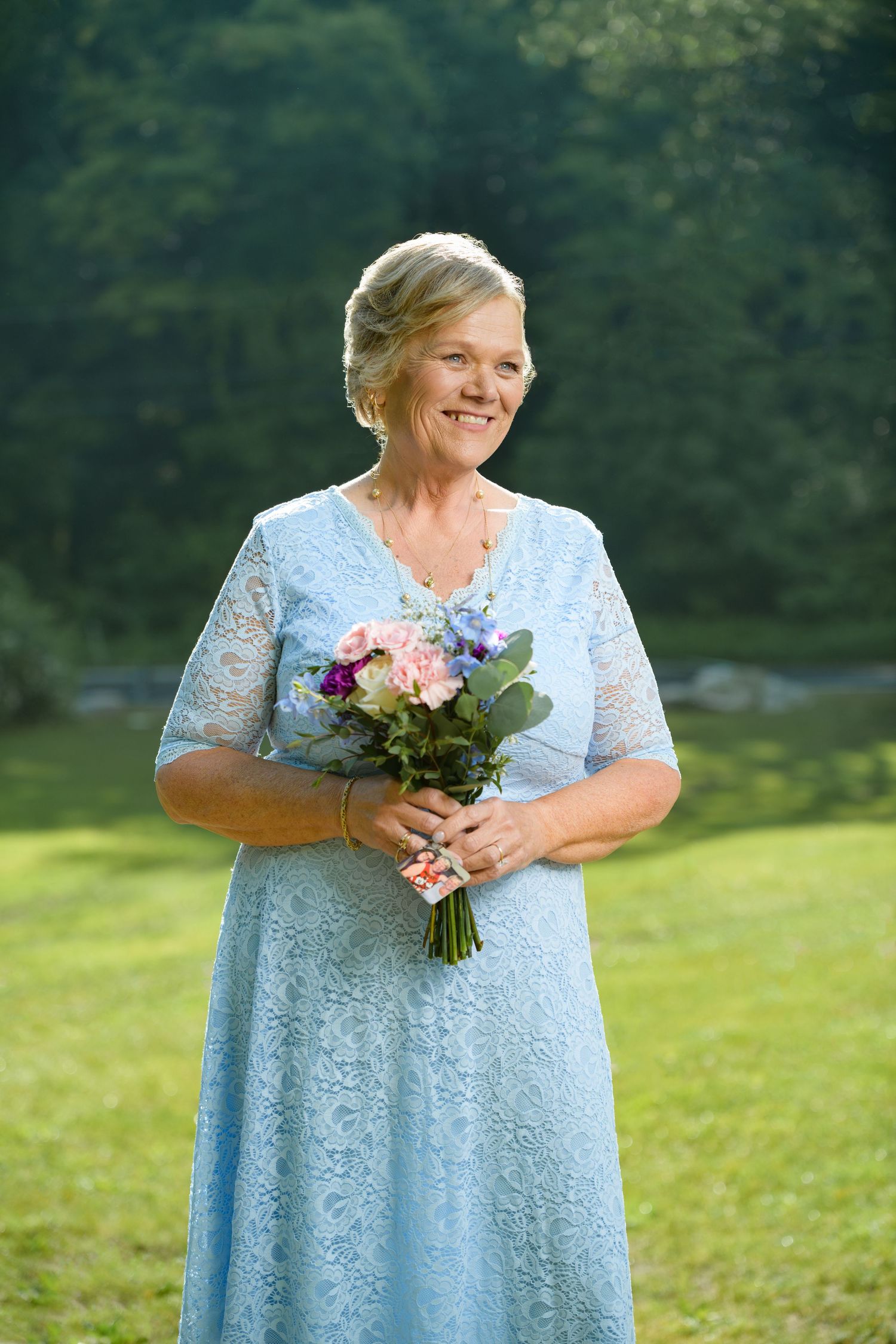An elegant older woman in a light blue lace dress holds a colorful bouquet while smiling outdoors in a garden.