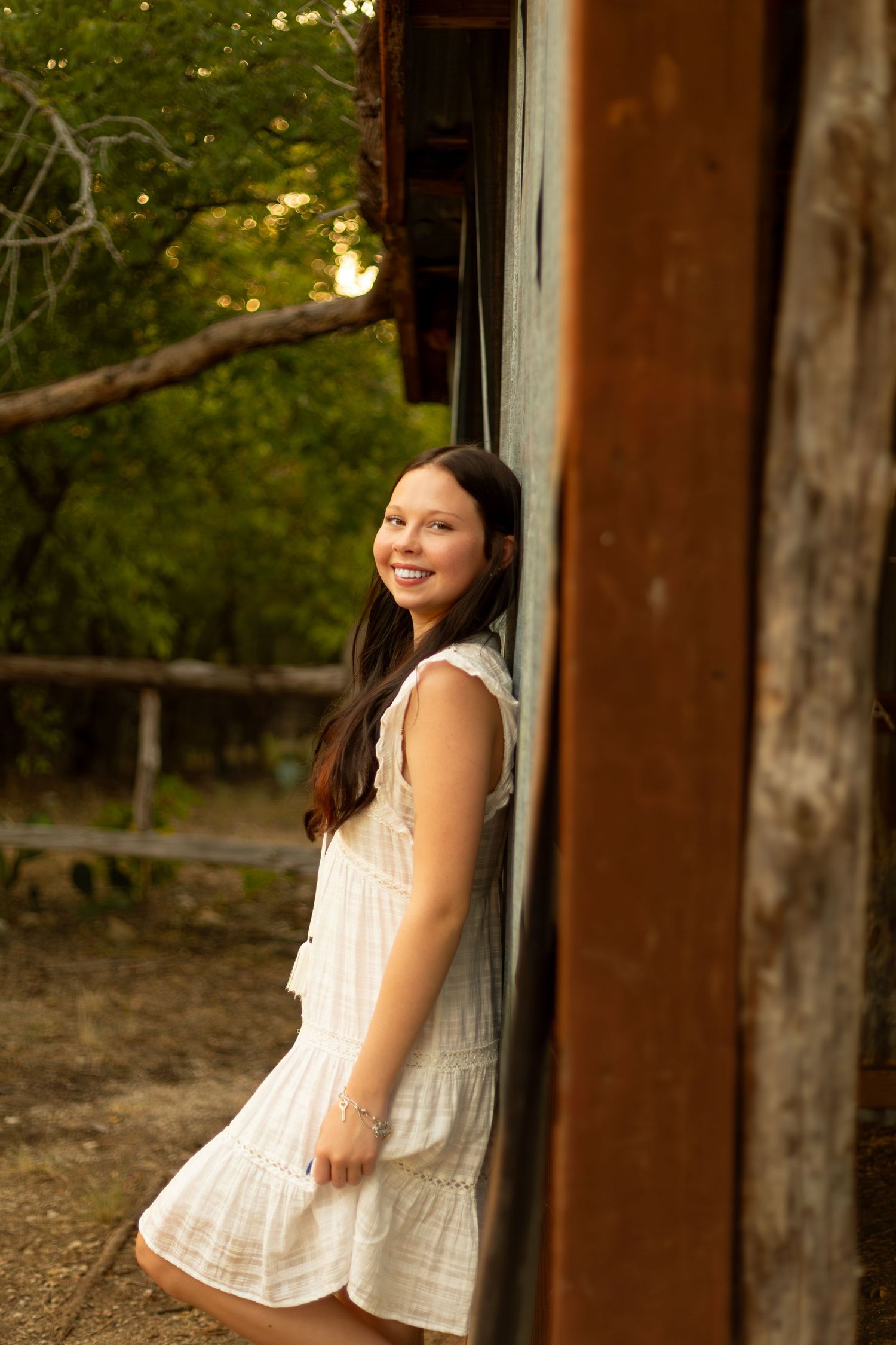A person in a white sleeveless top poses against wooden beams in an outdoor setting during golden hour.