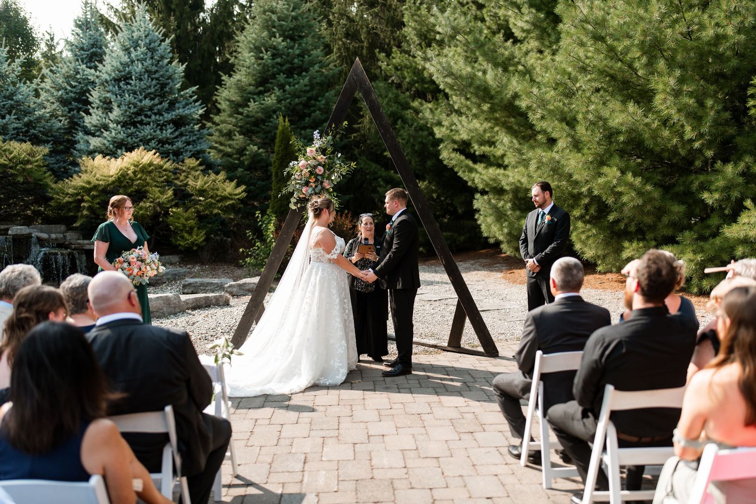 Wedding guests at Strarhroy Caradoc Sands Golf Club seated outdoors in white chairs among evergreen trees during a garden ceremony.