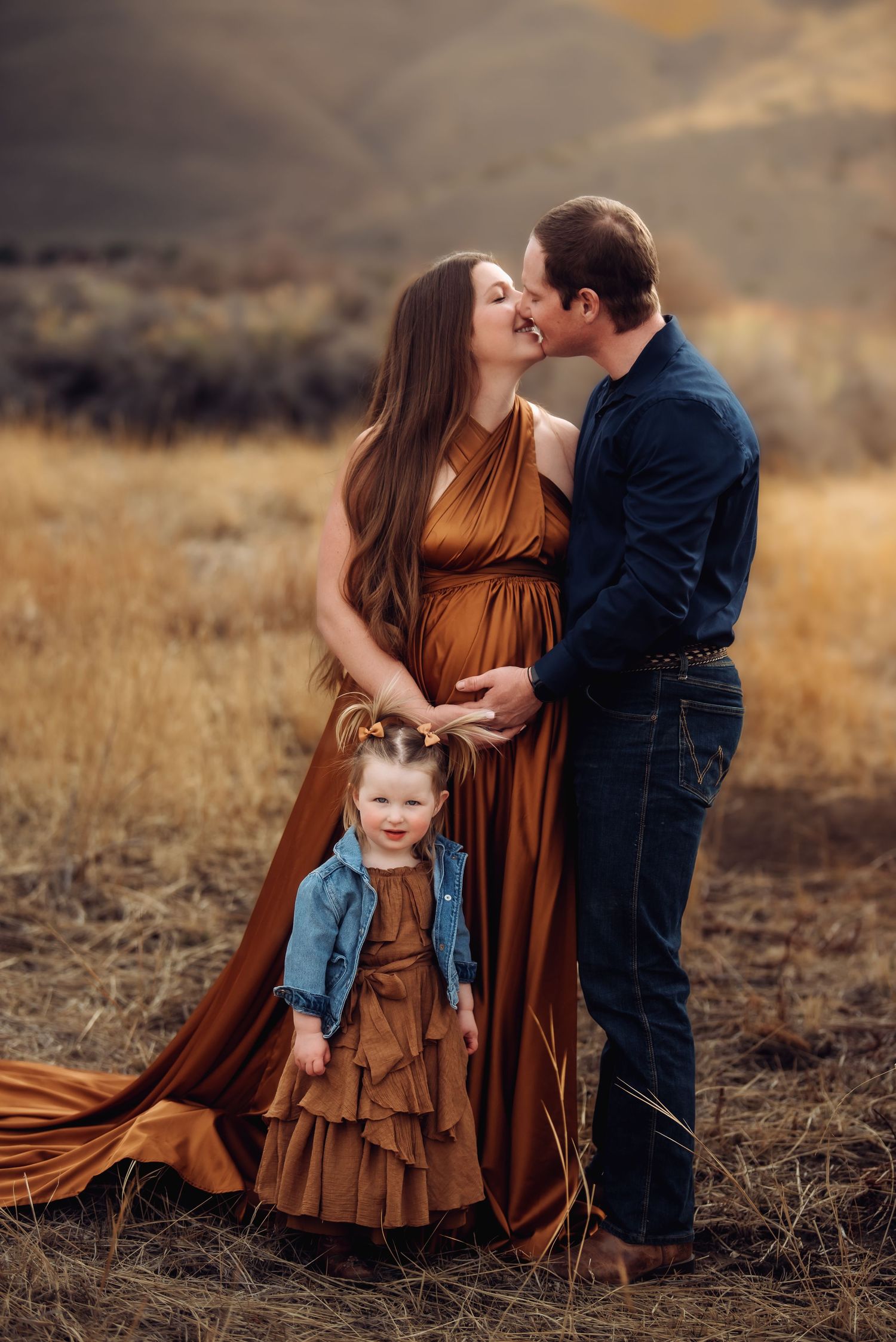 A family in coordinated brown and denim outfits embrace in a golden field during sunset.