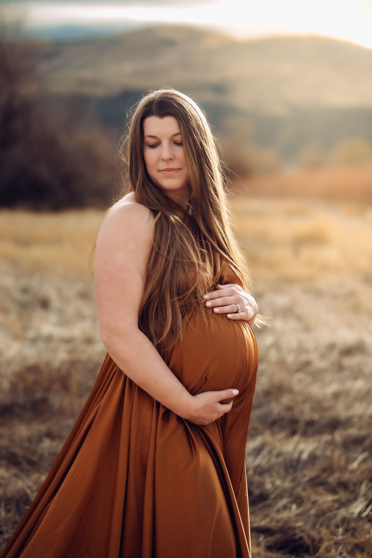 A woman in a flowing rust-colored dress poses gracefully in a golden field during sunset.