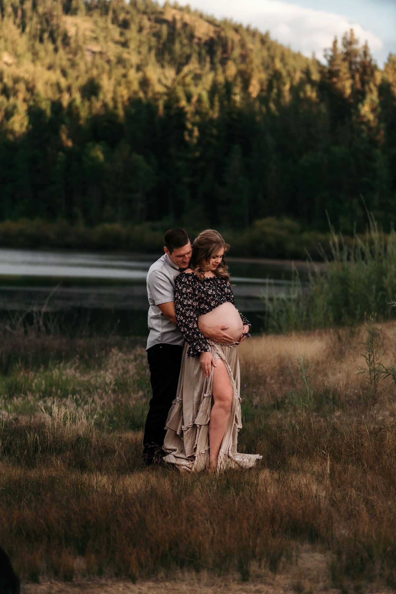 Romantic couple embraces by a serene lake at sunset with forest hills in the background.