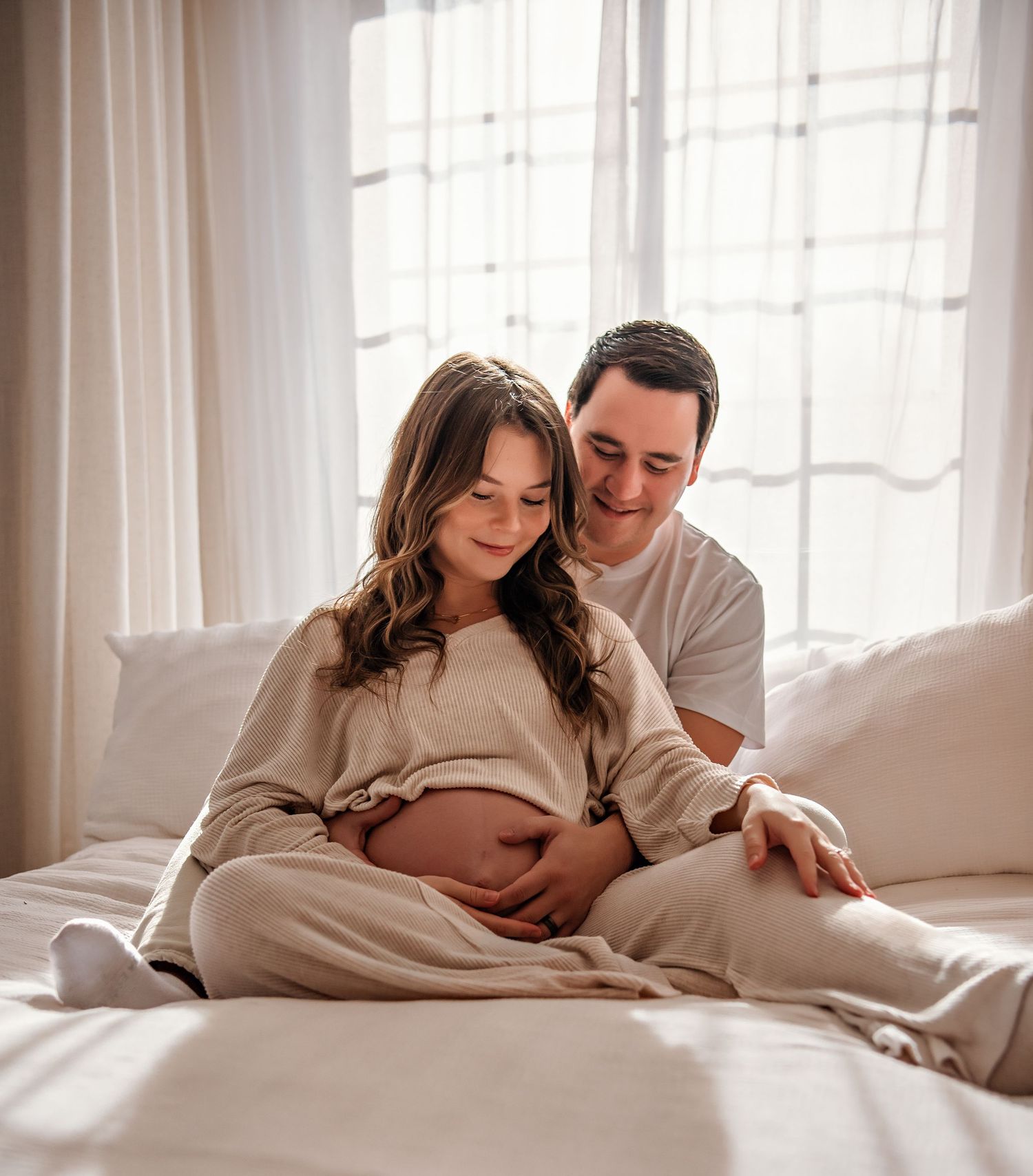 Couple relaxes together on white bedding in sunlit bedroom during intimate lifestyle maternity photoshoot.