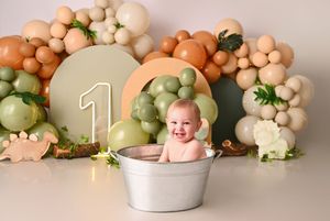 Smiling baby in silver tub at dinosaur-themed first birthday with green and tan balloon garland backdrop.