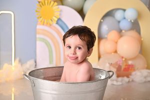 A smiling baby sits in a small metal tub during a colorful first birthday cake smash photo session.