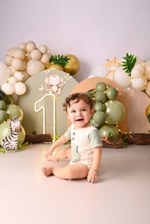 Smiling baby sitting cross-legged at a jungle-themed first birthday party with safari animal decorations and green balloons.