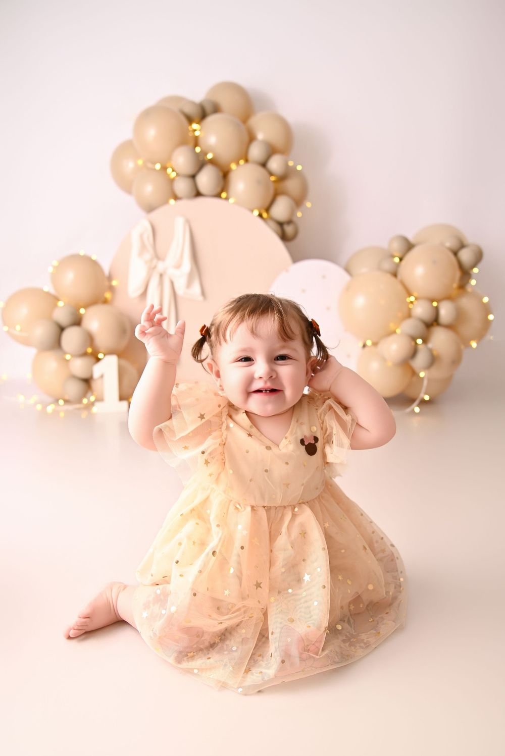 Smiling baby girl in gold dress sits at first birthday cake smash with beige balloon decorations.