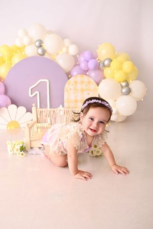 Smiling baby girl crawling at her first birthday cake smash with purple balloons and daisy decorations.