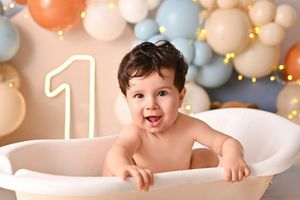 Baby boy smiling in a white tub surrounded by blue, orange, and beige balloons celebrating his first birthday.