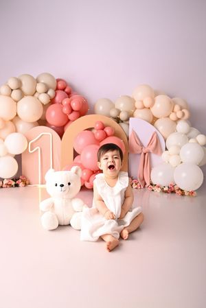 A baby girl in white dress sits beside a teddy bear at her first birthday with pink balloon decorations.
