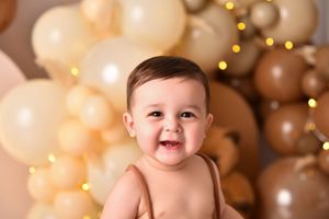 Baby sits against festive background with gold bokeh lights and peach colored balloons.