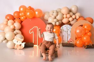 Baby in neutral-toned outfit sits among peach and orange balloon decorations for first birthday celebration.