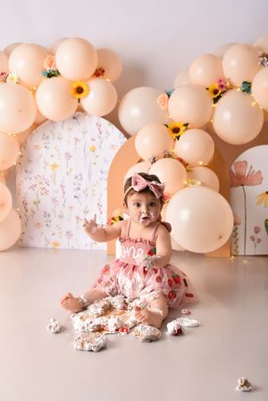 Baby wearing pink dress sits among peach-colored balloon arch and scattered cake during first birthday cake smash session.