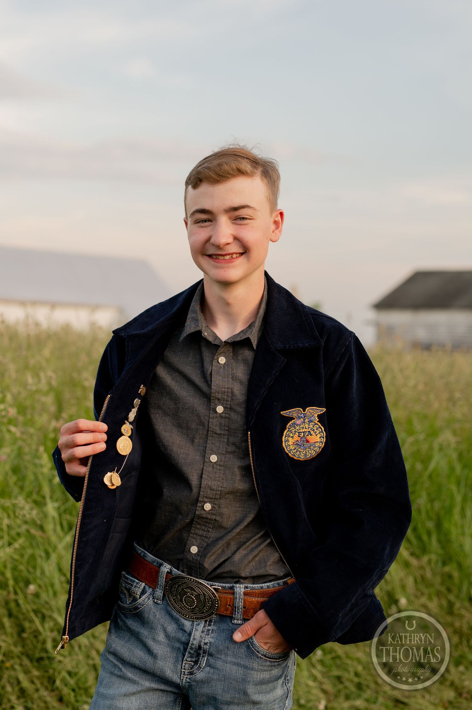 Student wearing FFA jacket and western belt poses in rural field setting.