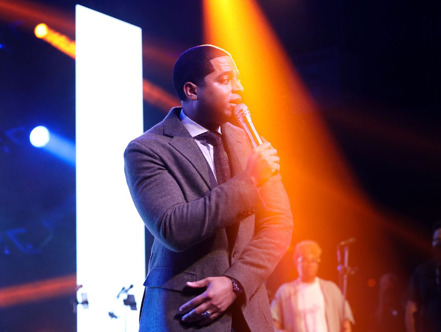 A performer in a dark suit holds a microphone on stage with dramatic orange and blue lighting effects during a show.