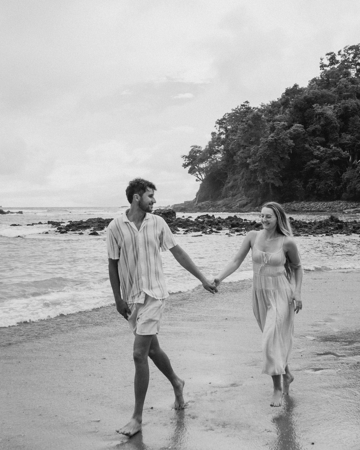 Black and white photo of a couple running together on the beach, with the man leading and both smiling.