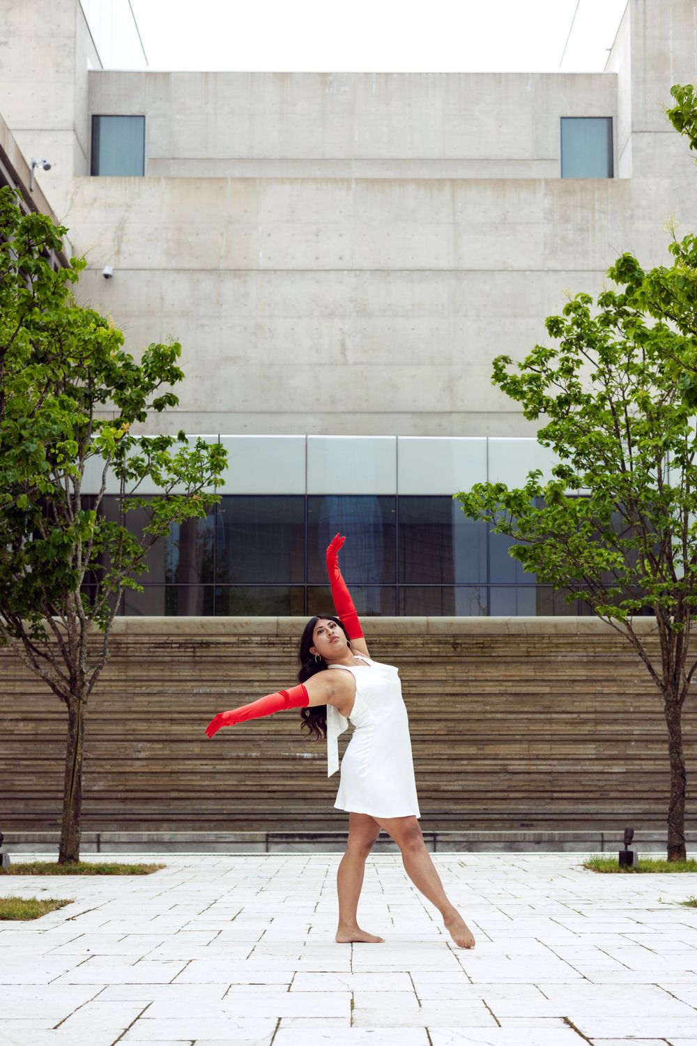 Dancer in white dress performs graceful pose with red fabric against modern building exterior.