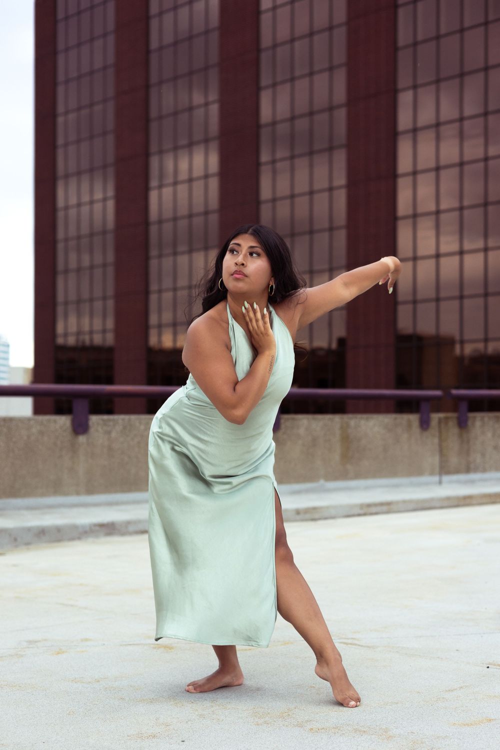 Dancer in mint green dress performs expressive movement against urban backdrop with glass windows.