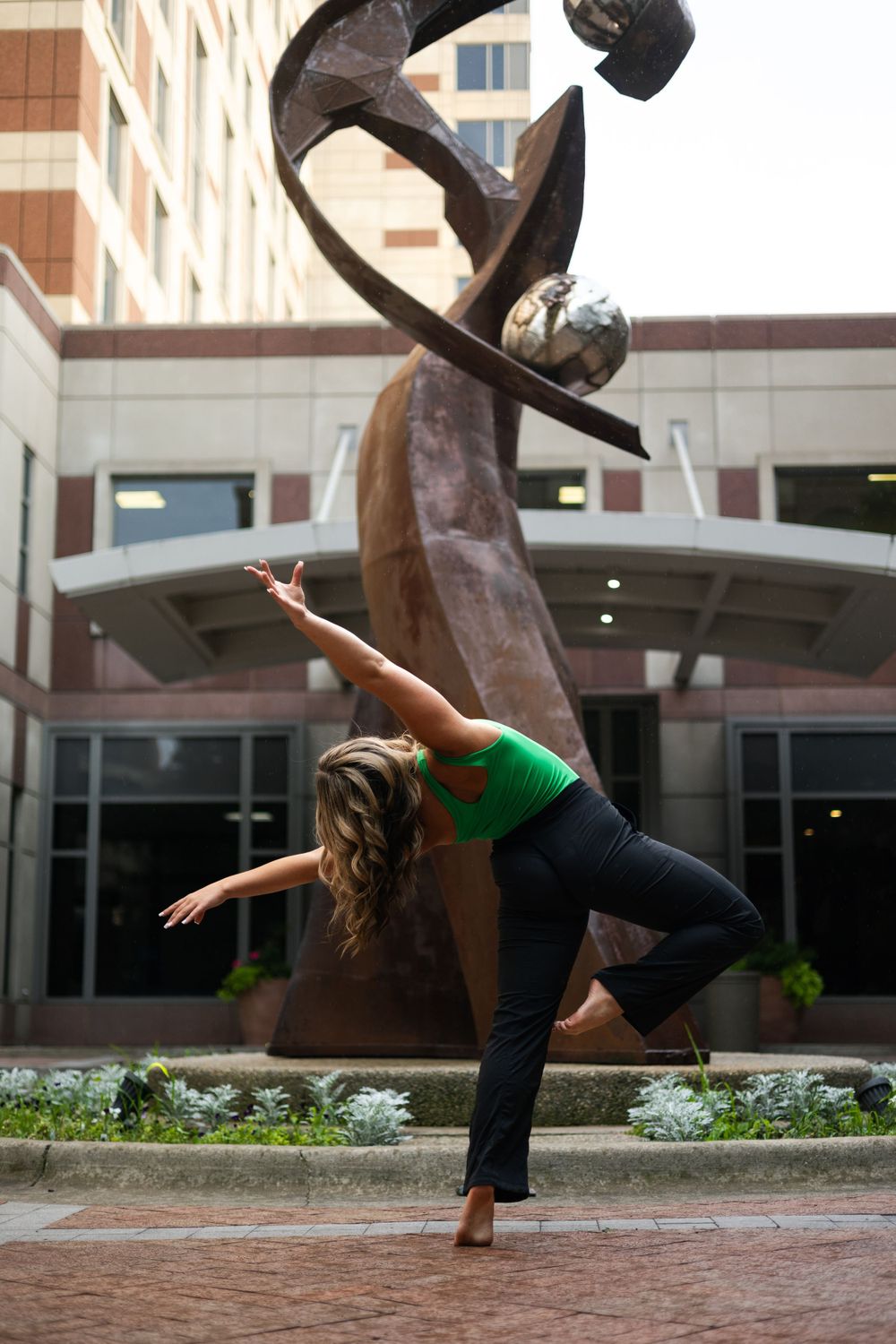 Dancer performs graceful pose in front of modern sculptural art installation at urban building entrance.