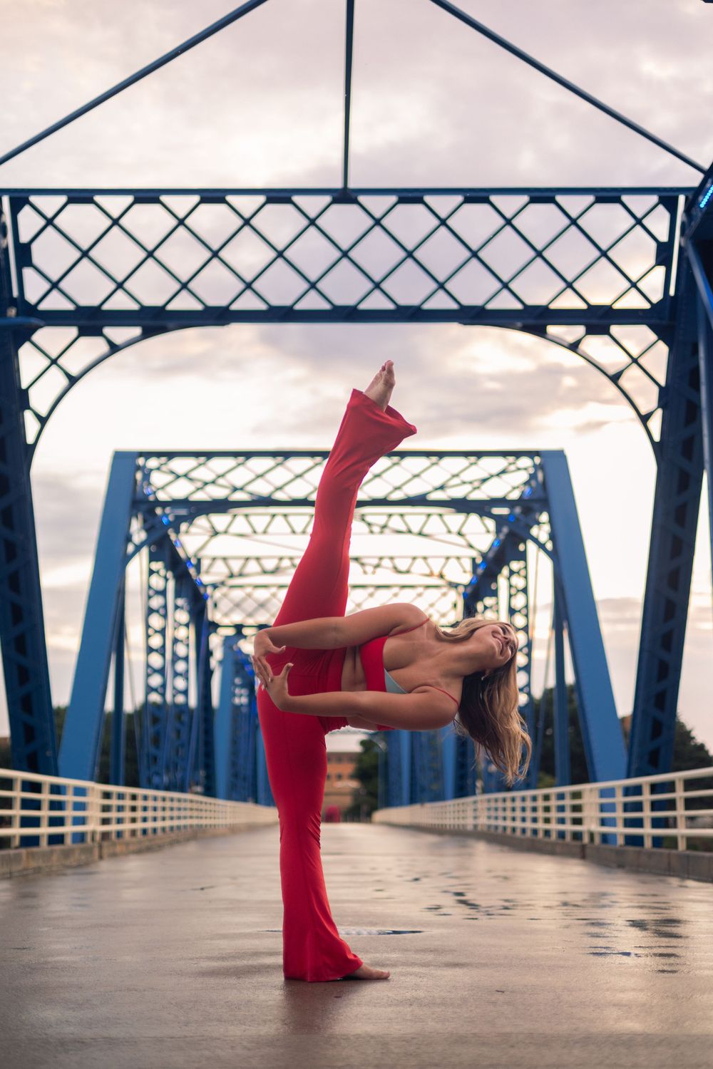 Graceful dancer in red performs a standing split on a blue steel bridge during an overcast day.