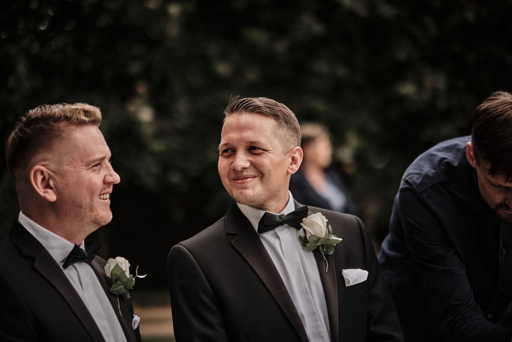 Formal wedding party members in black tuxedos with boutonnières share a moment during outdoor ceremony.