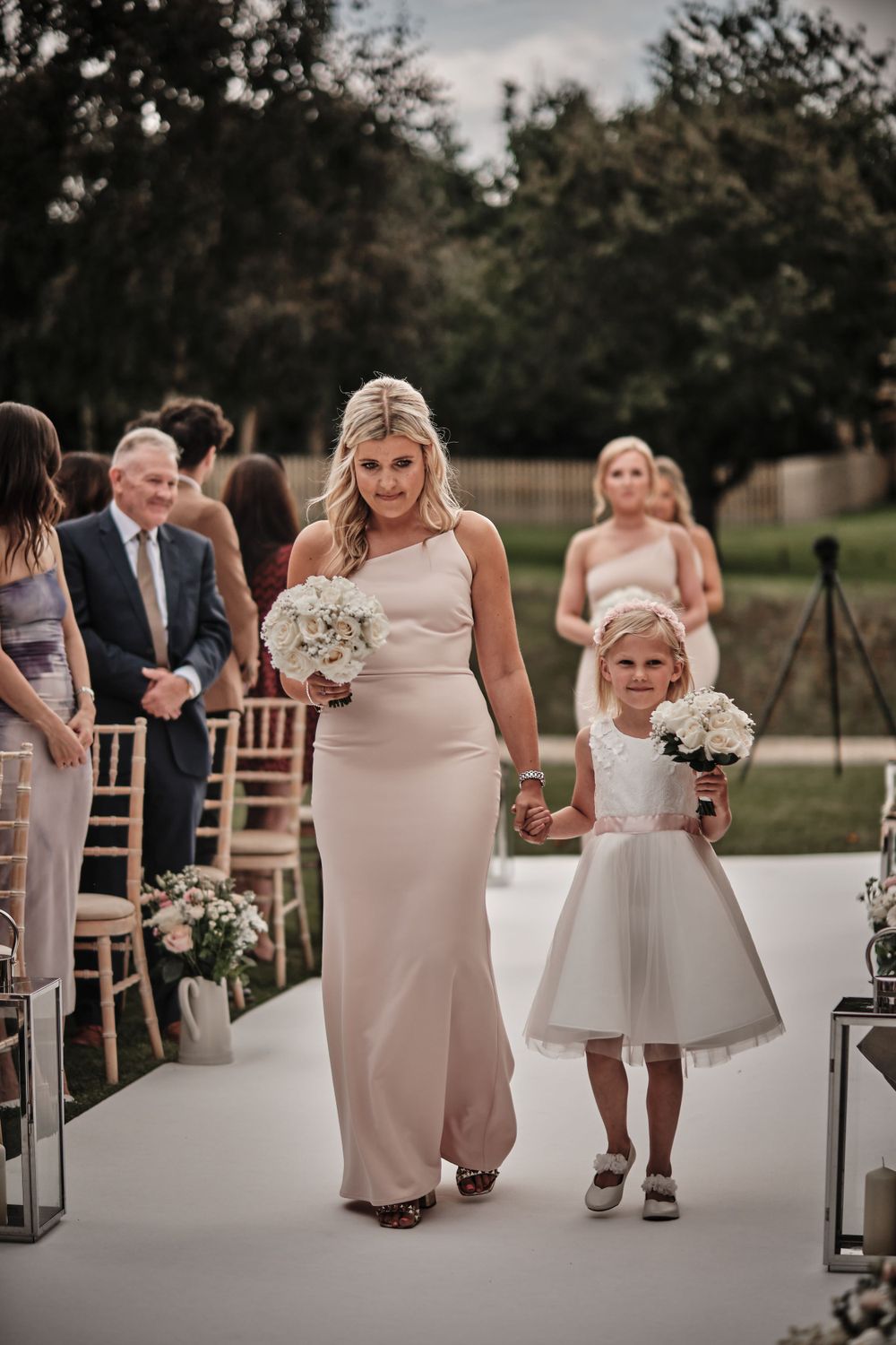Wedding processional on white aisle runner with attendants in blush gowns carrying white bouquets.