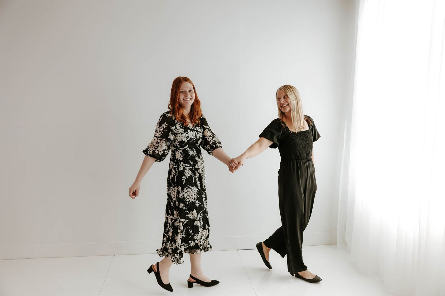 Two friends sit back to back wearing casual sweaters and jeans against a white wall background for branding photos.