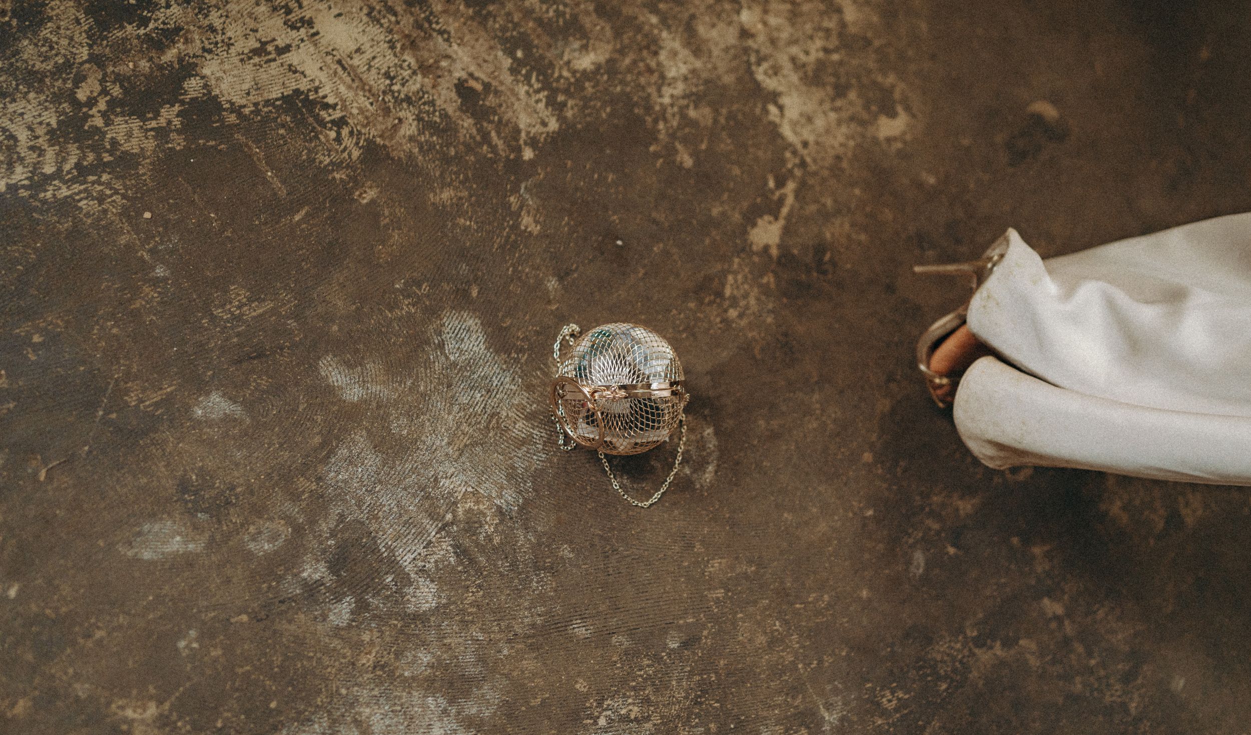 Gold coloured spherical handbag on a concrete floor with foot of guests in view. Editorial wedding