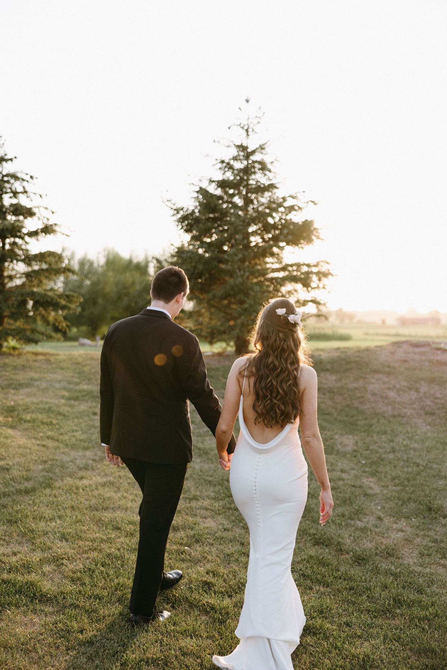 Couple walks hand in hand through field at sunset in wedding attire.