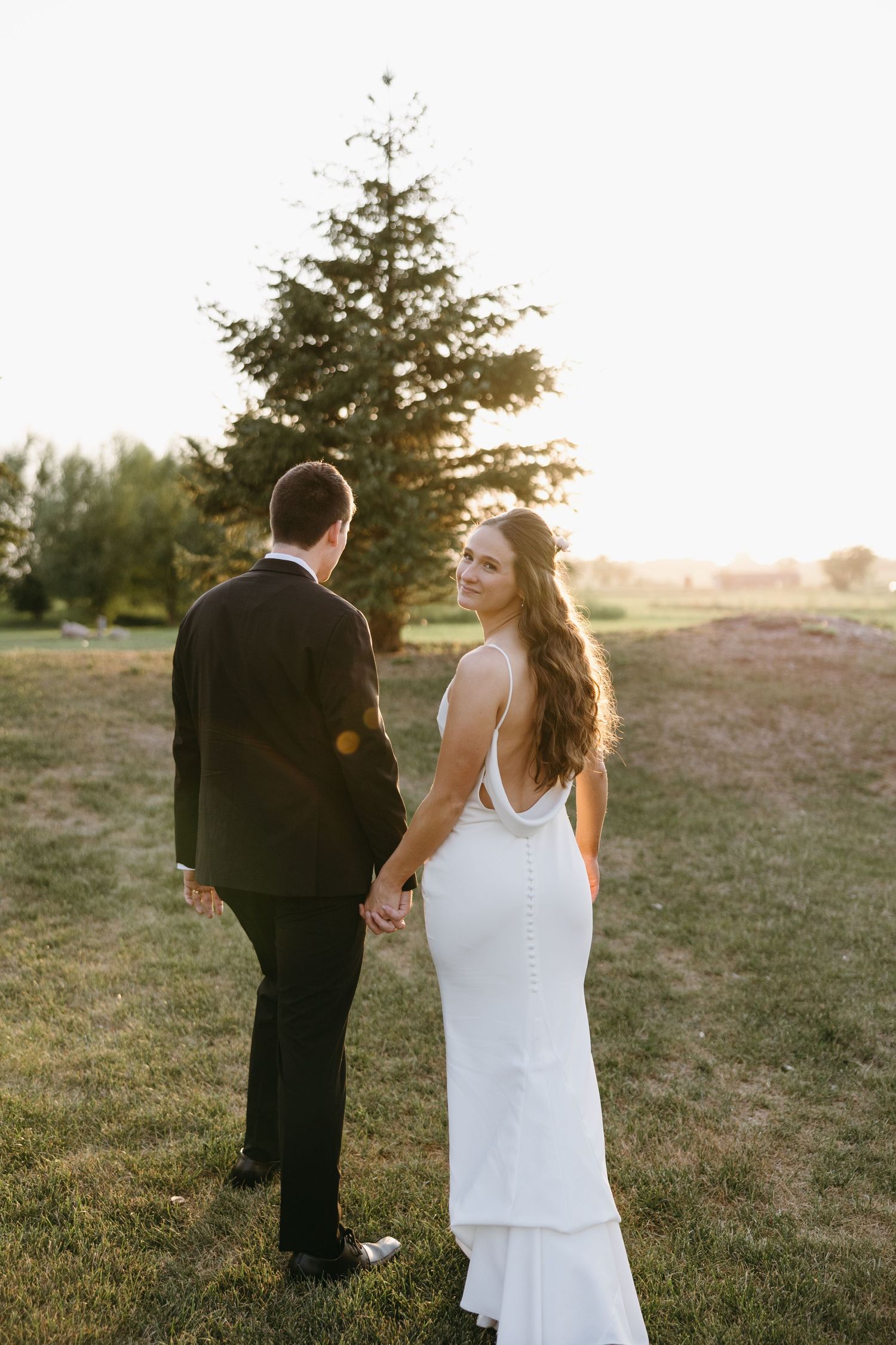 Couple walking away from camera through grassy field at golden hour.